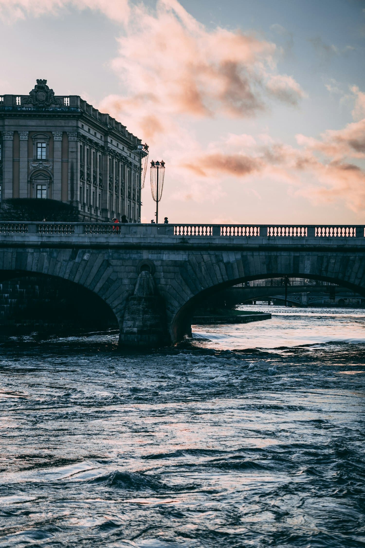 A stone bridge in central Stockholm at dusk, with water rushing beneath and the Parliament House behind