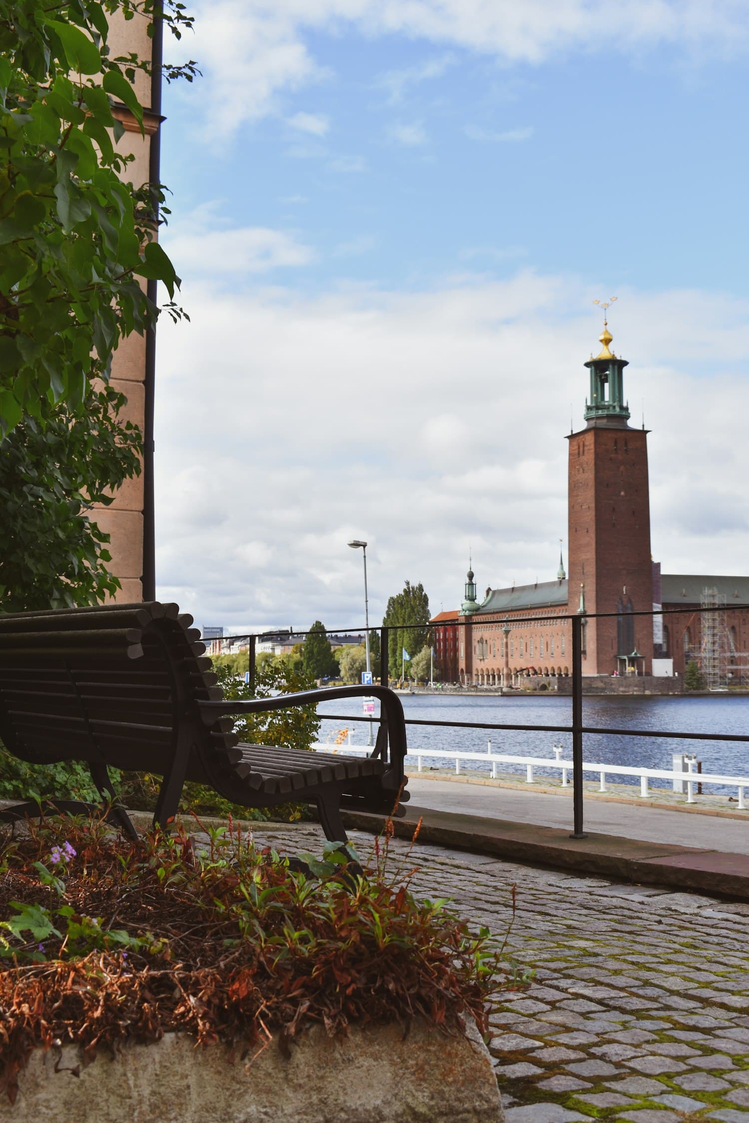 The brick tower of Stockholm City Hall seen from the waterside promenade, with a bench beneath autumn trees